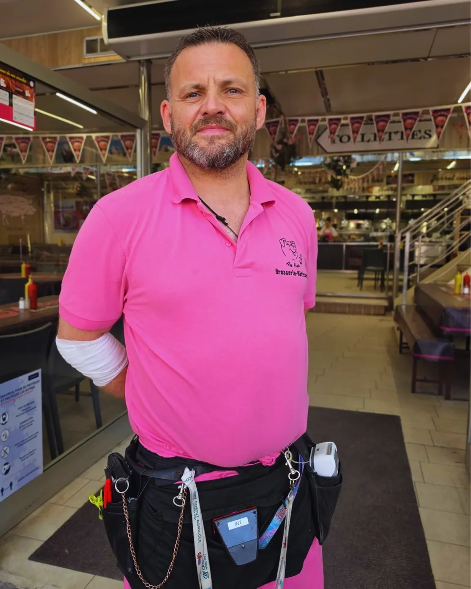 Portrait souriant d'un serveur en polo rose de l'enseigne devant l'entrée décorée de fanions.