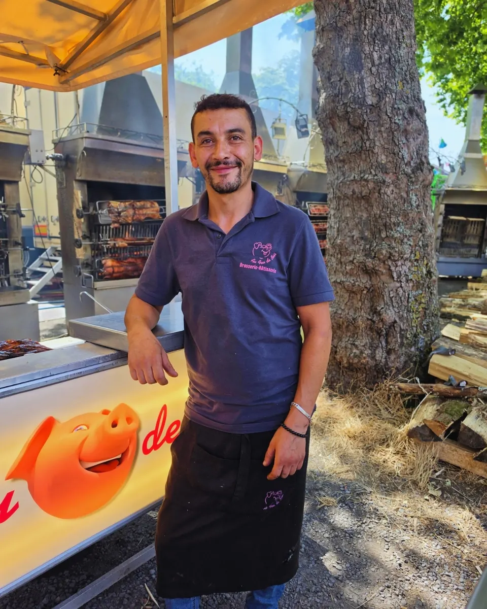 Rôtisseur souriant accoudé au comptoir orné du grand cochon rieur, rôtissoires et bois de chauffe derrière lui.