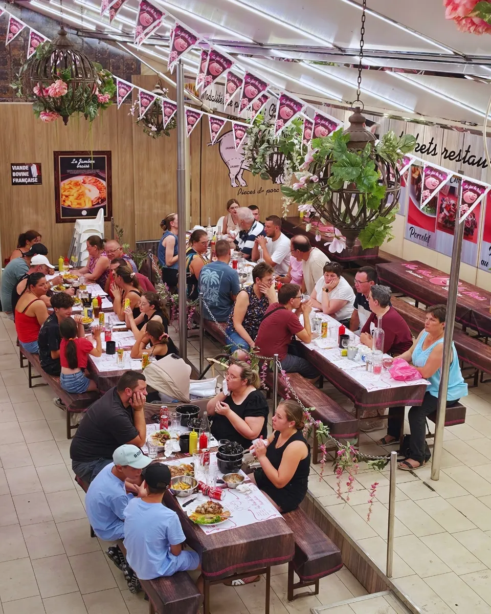 Vue en plongée sur deux longues tablées de la salle où des familles déjeunent sous des guirlandes de fanions roses au cochon