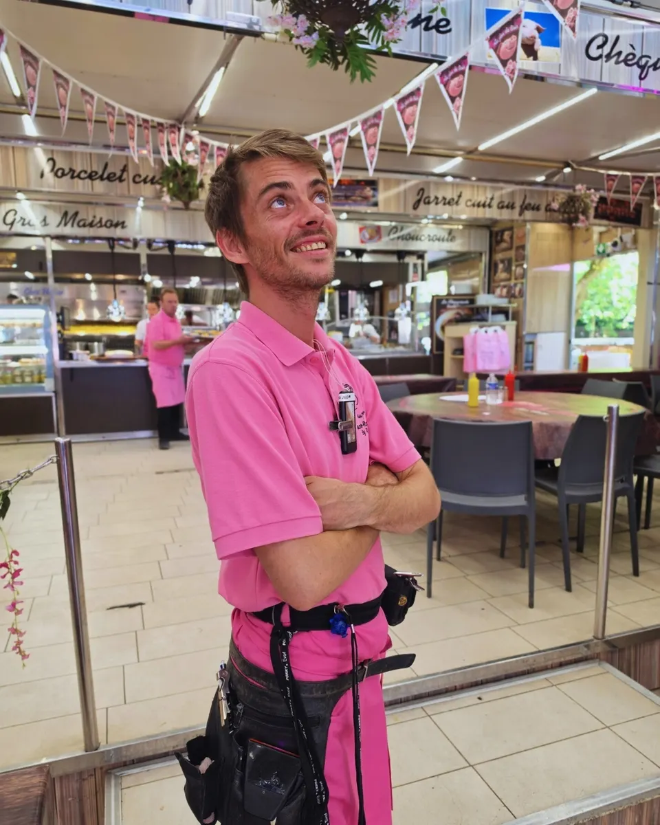 Jeune serveur en polo rose posant bras croisés dans la salle décorée de fanions au cochon.