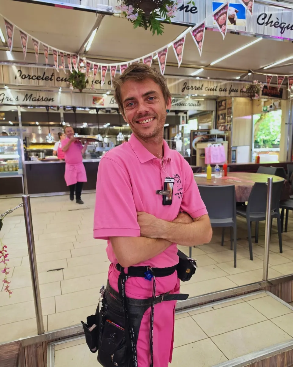 Serveur au grand sourire en polo rose brodé du cochon, dans la salle du stand.