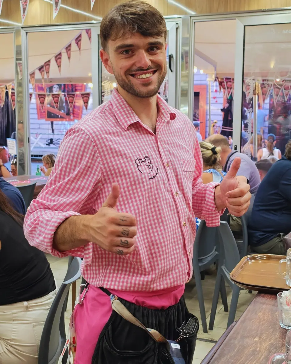 Jeune serveur enthousiaste, pouces levés, en chemise vichy rose au logo cochon devant la salle comble.