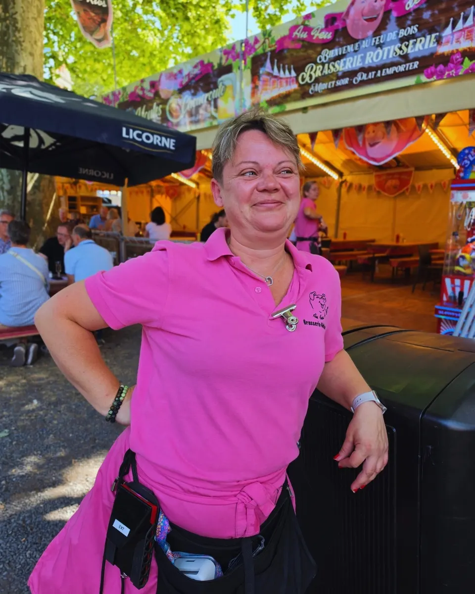 Serveuse souriante devant la bannière Bienvenue Au Feu de Bois - Brasserie Rôtisserie et son cochon rose.