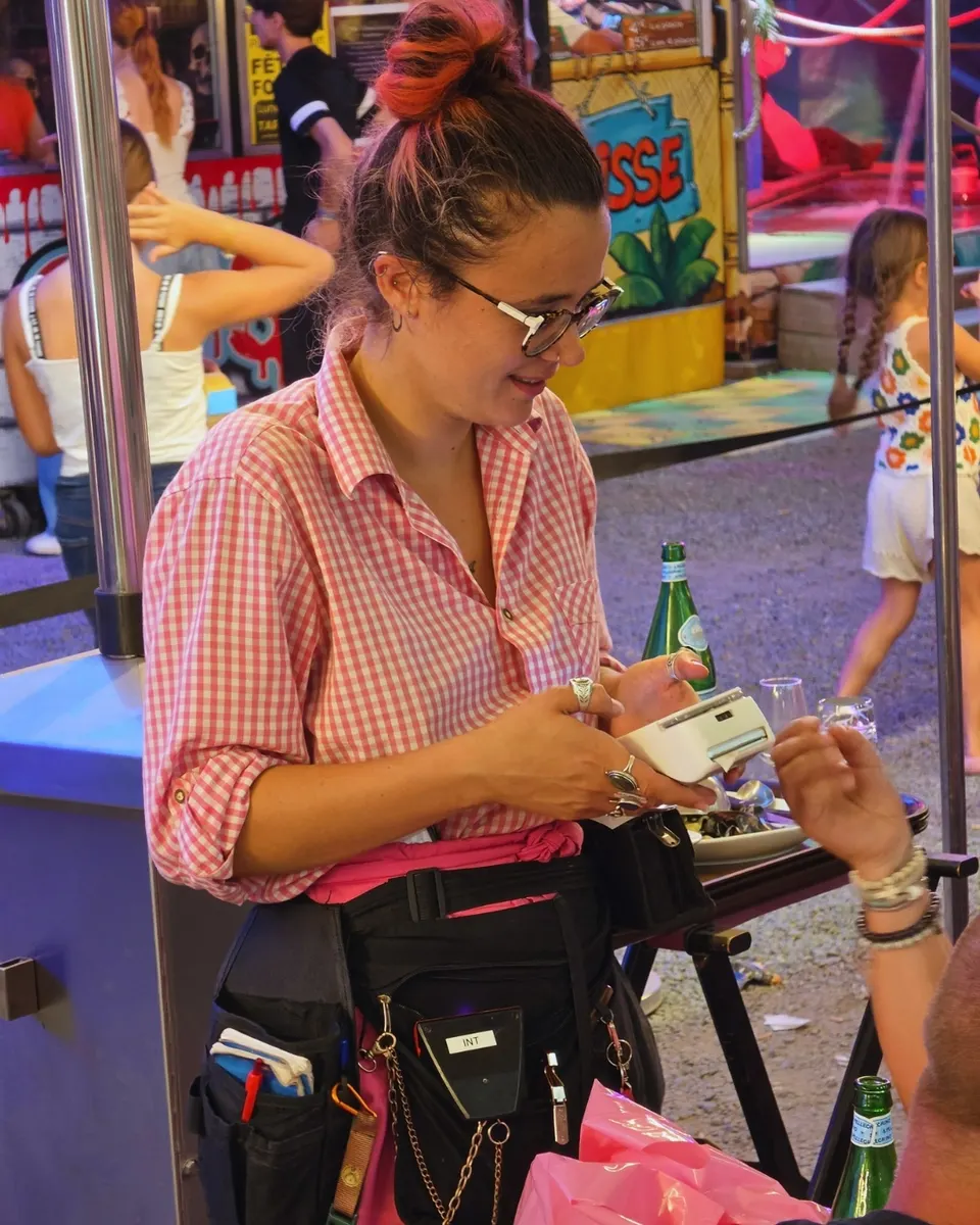 Serveuse en chemise vichy rose encaissant un client à la nuit tombée, lumières de la fête foraine.