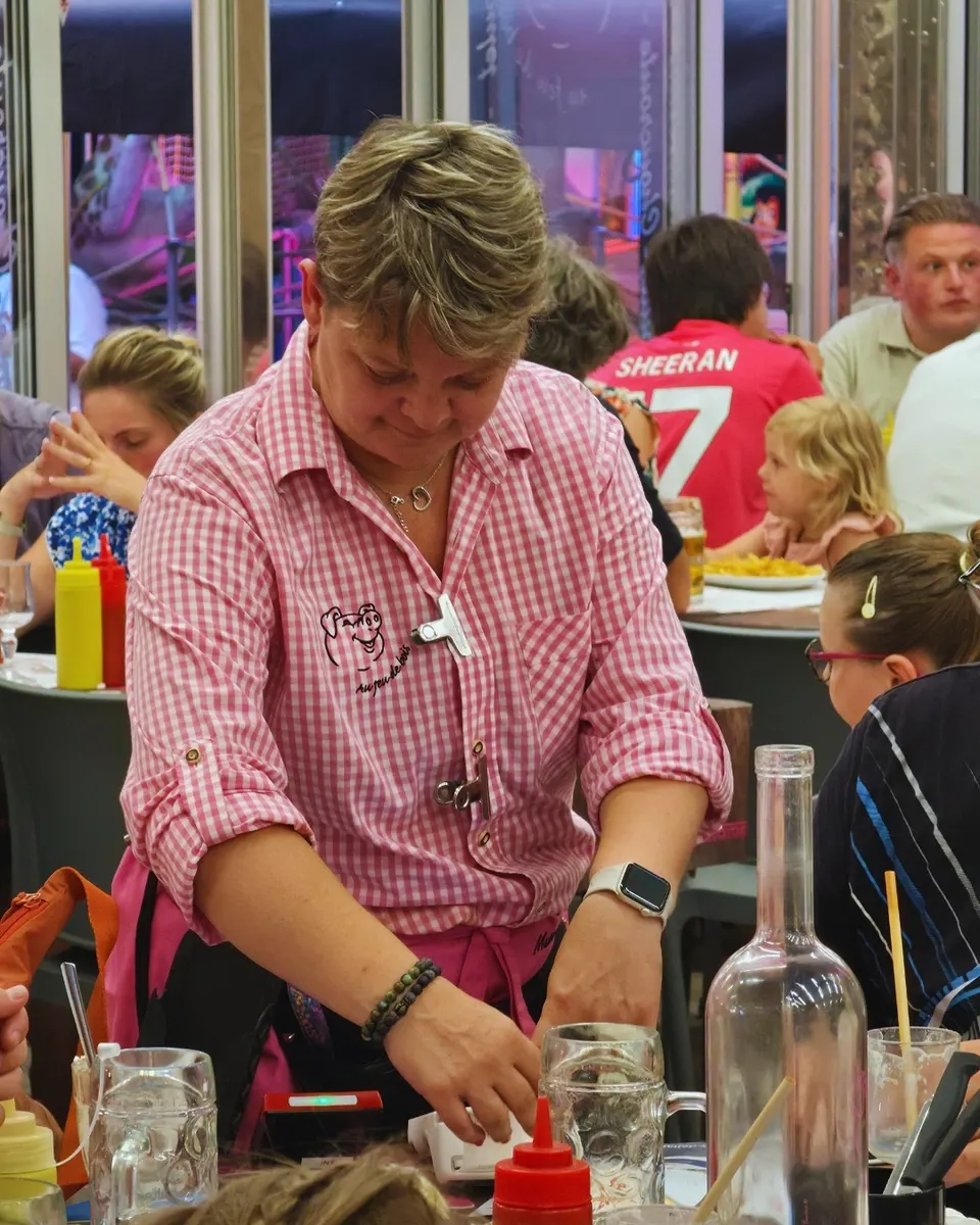 Serveuse en uniforme vichy rose encaissant un client au terminal, au milieu des tables animées en soirée.