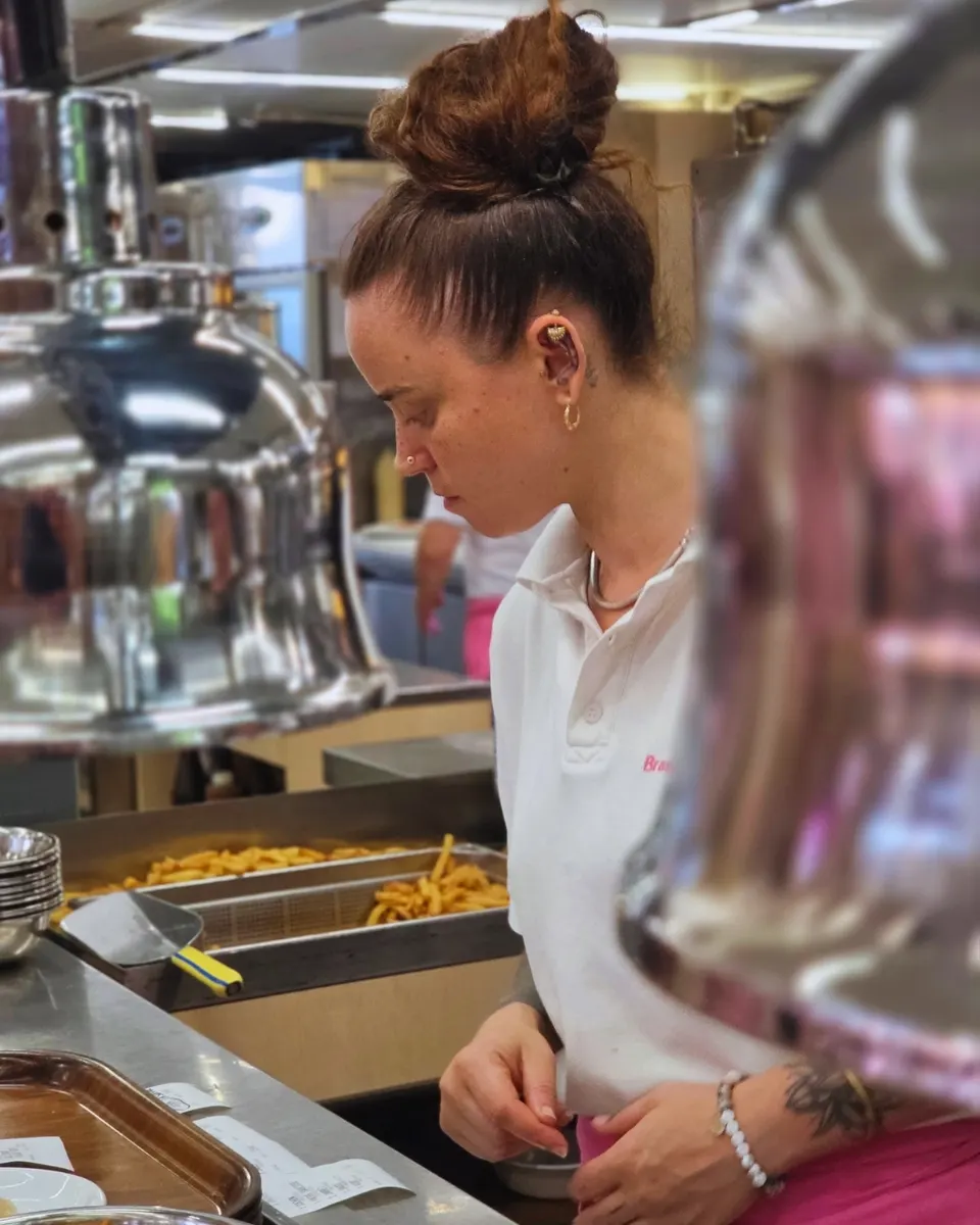 Serveuse concentrée au poste des frites derrière le comptoir inox de la cuisine.