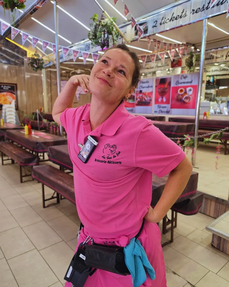 Serveuse espiègle en polo rose dans la salle aux longues tables-bancs en bois.