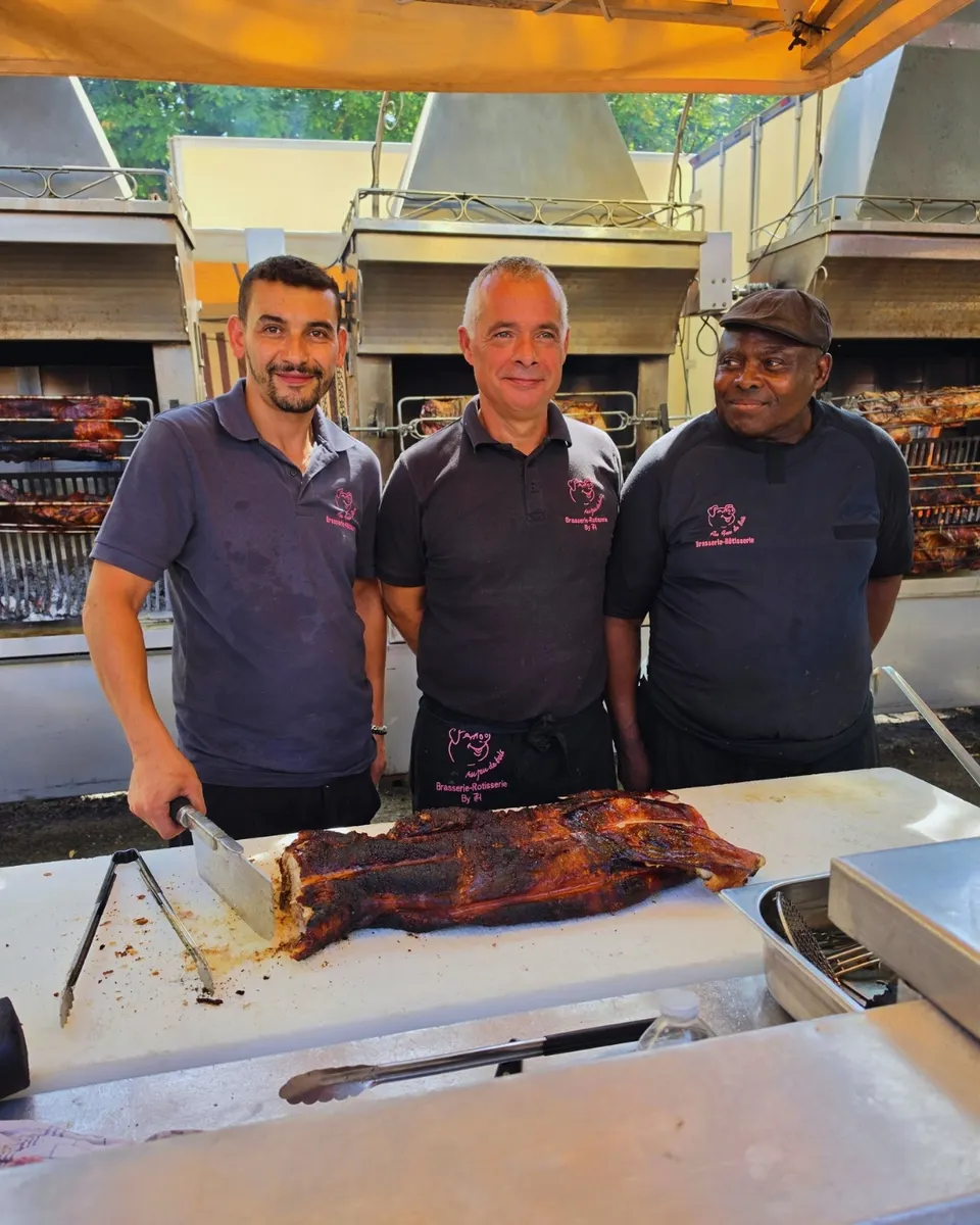 Les trois rôtisseurs souriants derrière un cochon entier rôti, rôtissoires à broches en arrière-plan.